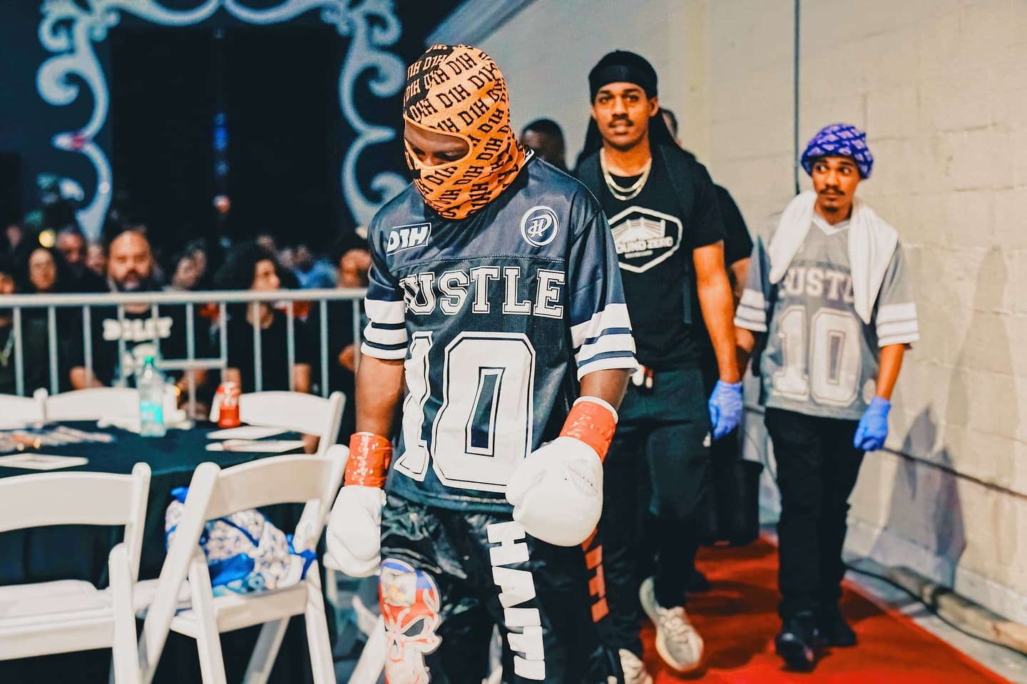 Kenneth Sene walking out to the ring in a mask and a black HUSTLE 10 jersey, his team behind him